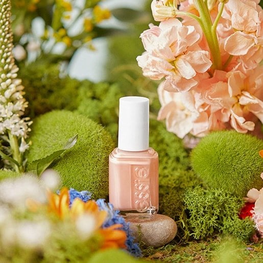 A bottle of pink nail polish on a small stone and surrounded by flowers and moss-covered stones