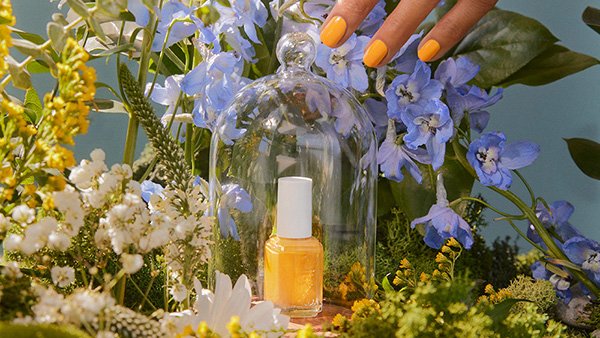  A hand reaching to lift a clear glass cloche that contains a bottle of nail polish surrounded by flowers
