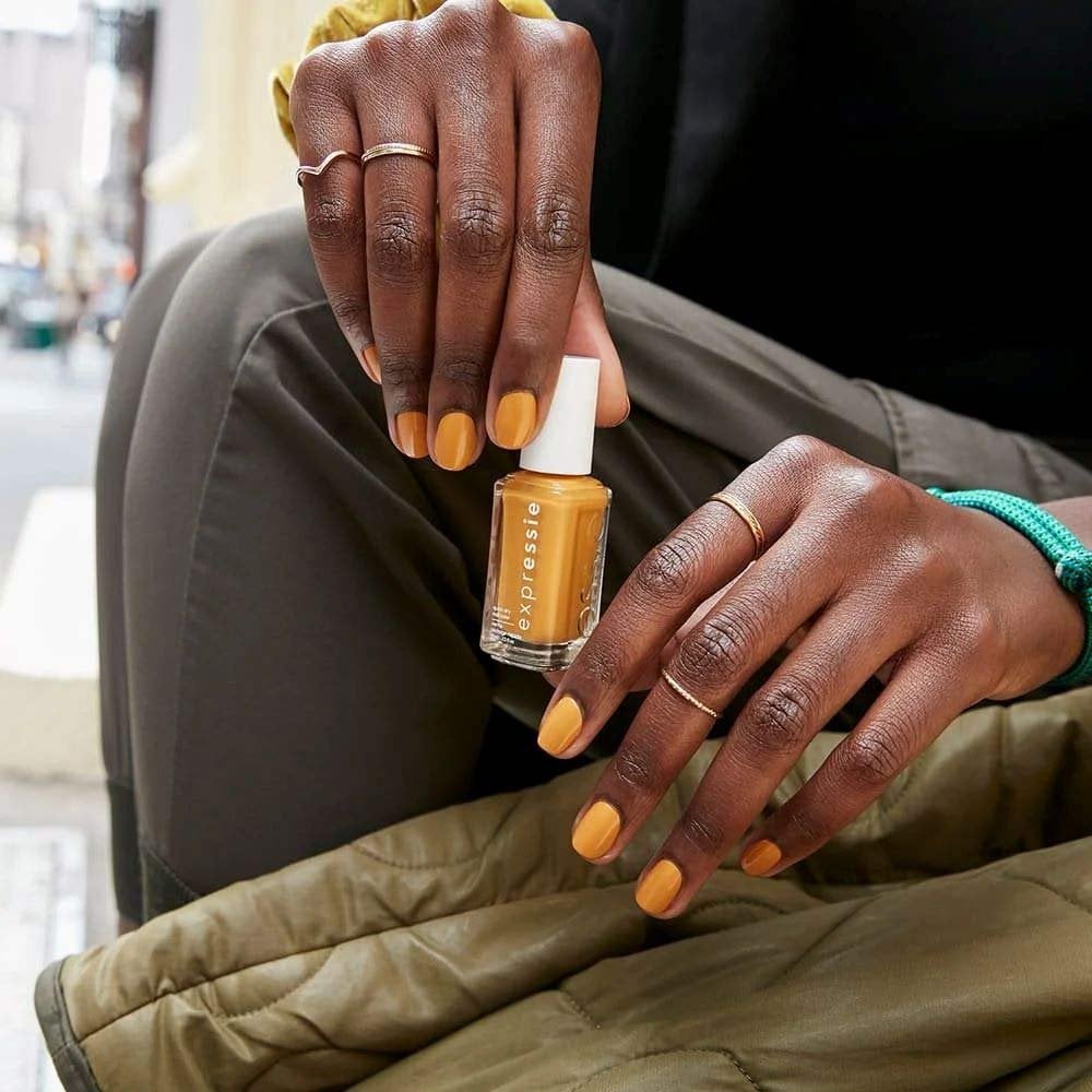 Model with dark skin and orange nails painted with essie nail polish sitting on a city street